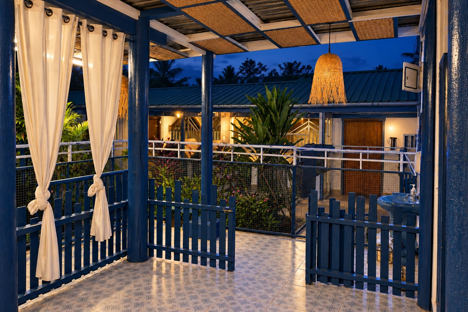 The covered deck at night: white curtains tied to the blue posts under a woven pendant lamp