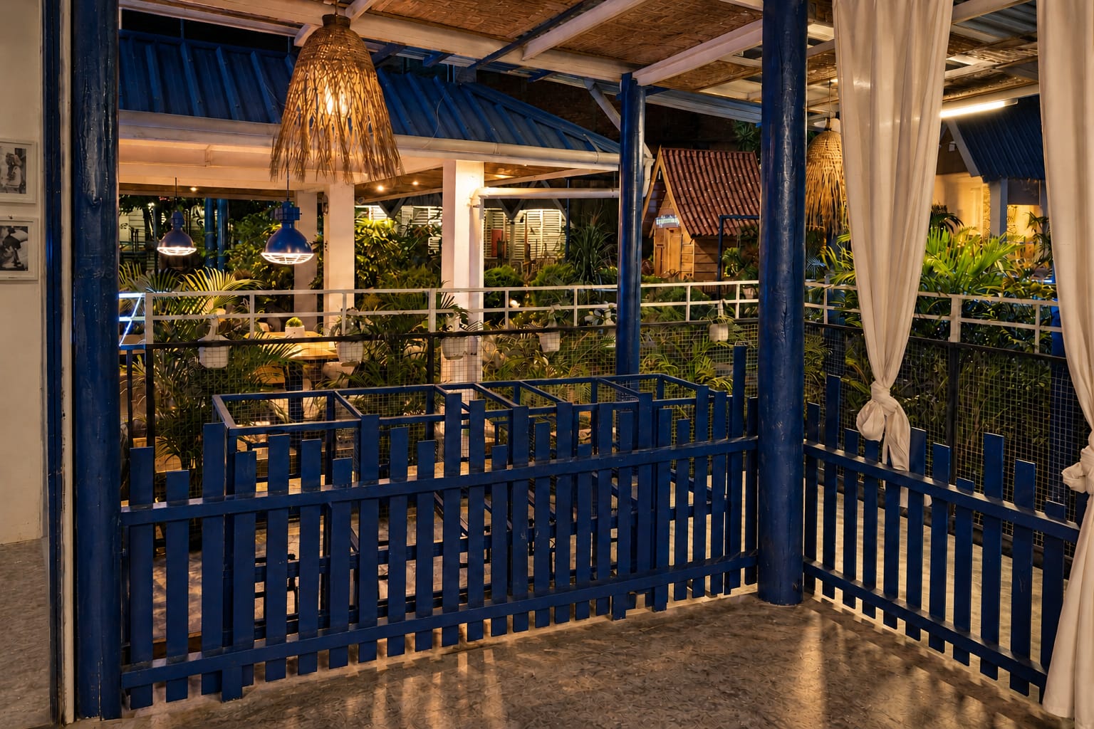 The deck at dusk: cobalt railings, tied white curtains and a woven lamp, with the garden beyond